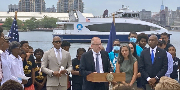 Group of people stand outdoors by an NYC Ferry waterfront, with a boat in the background. A suited Andrew Kimball speaks at a podium flanked by officials and attendees; flags are visible on either side, and Mayor Eric Adams is standing on the left of Kimball.