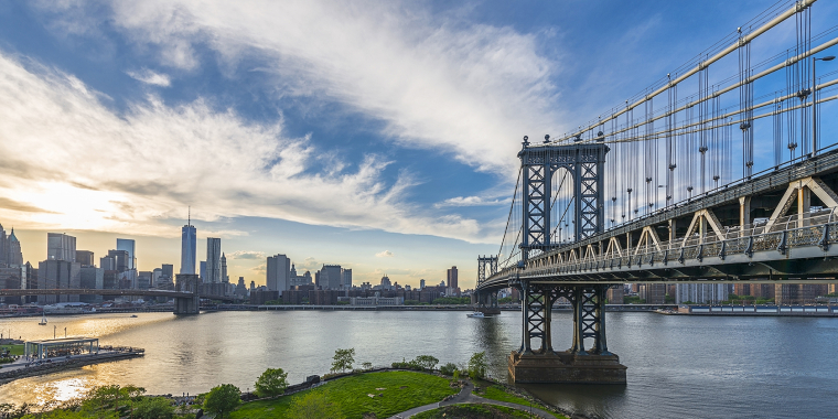 The Manhattan Bridge spans the East River with the New York City skyline and skyscrapers in the background under a partly cloudy sky at dawn. Lush greenery and waterfront paths are visible in the foreground.