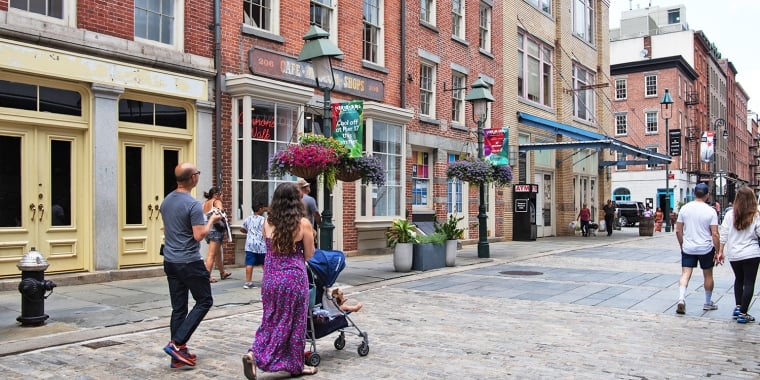 People stroll in South Street Seaport neighborhood on a cobblestone street lined with historic brick buildings. A woman pushes a stroller, and colorful hanging flower baskets adorn the scene. Banners and vintage-style lamps add charm under a partly cloudy sky.