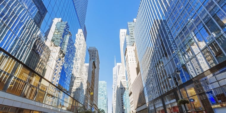 A vibrant city street flanked by tall skyscrapers with mirrored glass. The road is busy with vehicles, including cars and trucks, and pedestrians on the sidewalks. The sky is clear and blue, reflecting off the buildings.