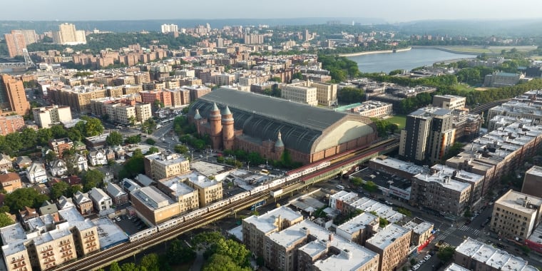 Aerial rendering of Kingsbridge Armory, a large brick building with a curved glass roof, surrounded by residential and commercial buildings. A train is visible on tracks in the foreground, with greenery and a body of water in the background.