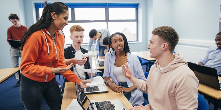 A group of college students in a classroom are collaborating, smiling, and discussing while using laptops. One student stands, holding a 3D-printed wind turbine model, engaging with seated classmates in a bright, modern room.