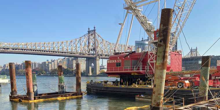Construction site of Offshore Wind NYC Waterfront Pathways Program, showing a red crane barge on a river with the Queensboro Bridge in the background. Several pilings are in the water, and buildings line the opposite riverbank under a clear blue sky.