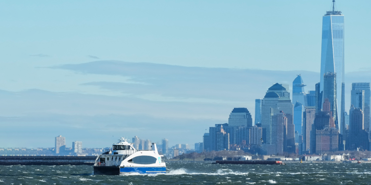 White and blue NYC Ferry travels across choppy water with the New York City skyline, including the One World Trade Center, visible in the background under a clear sky.