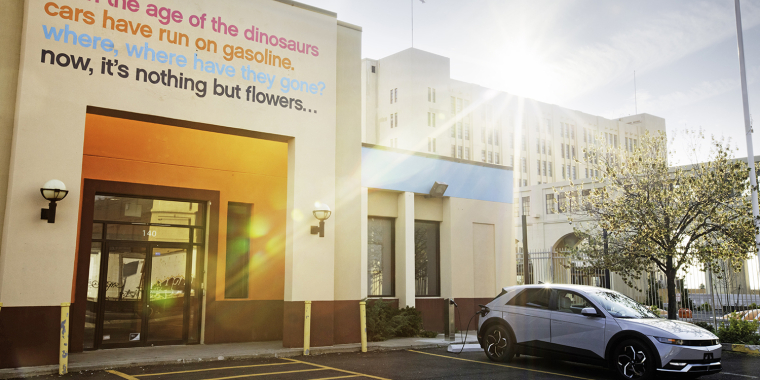 A silver electric car charging while parked at a curb outside a building with text on the wall referencing dinosaurs, gasoline, and flowers. The sun shines brightly, casting long shadows, and a large government building is visible in the background. The charging port is by itselectric, a Brooklyn-based electric vehicle curbside charging company.