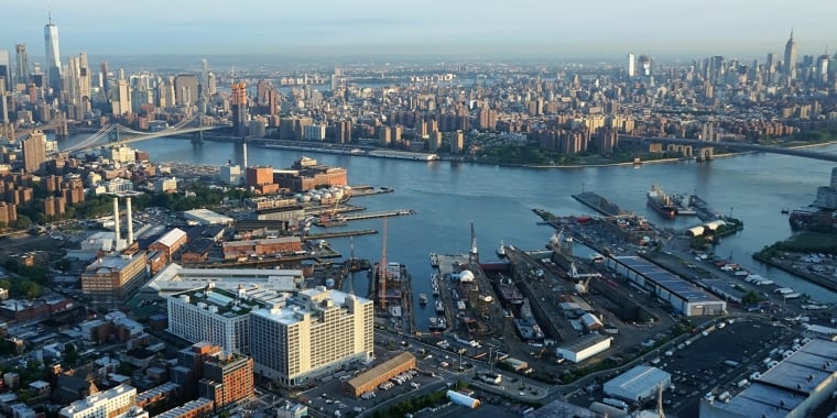 Aerial view of Brooklyn Navy Yard with piers, warehouses, and streets in the foreground, and the East River separating Brooklyn from the Manhattan skyline in the background, including several tall skyscrapers. Photo by Jerry Trudell.