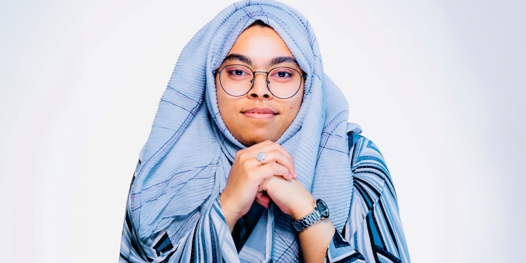 A female Cyber NYC intern, Samira Akram, wearing glasses and a light gray hijab, resting her chin on her hands. She is wearing a striped outfit and a watch, and is looking directly at the camera with a neutral expression against a plain background.
