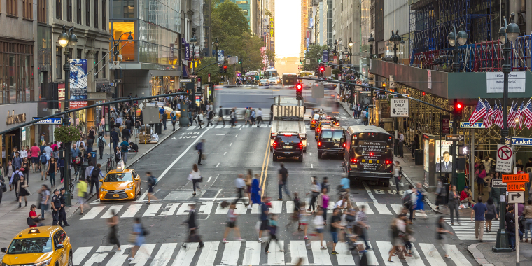 A busy 42nd Street in Midtown Manhattan, New York City, with many people crossing at crosswalks, yellow taxis, cars, buses, and tall buildings on both sides. Traffic lights and storefronts are visible, and the scene appears lively and bustling.