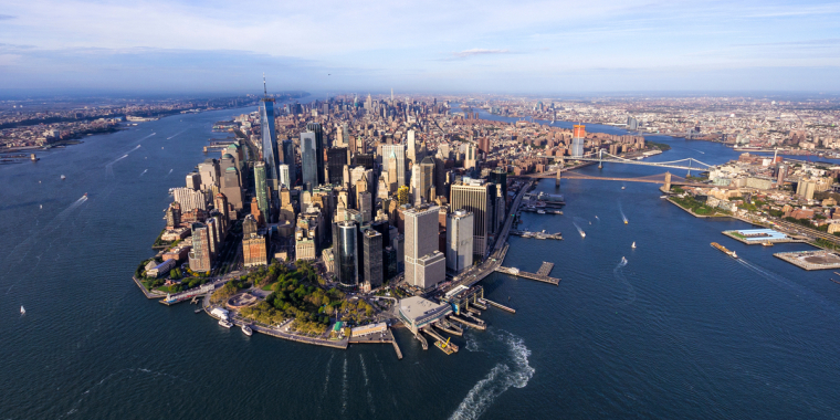 Aerial view of Lower Manhattan, New York City, surrounded by water with skyscrapers, ferries, and bridges visible, including the Brooklyn Bridge, under a clear blue sky. Photo courtesy of Eloi Omella.