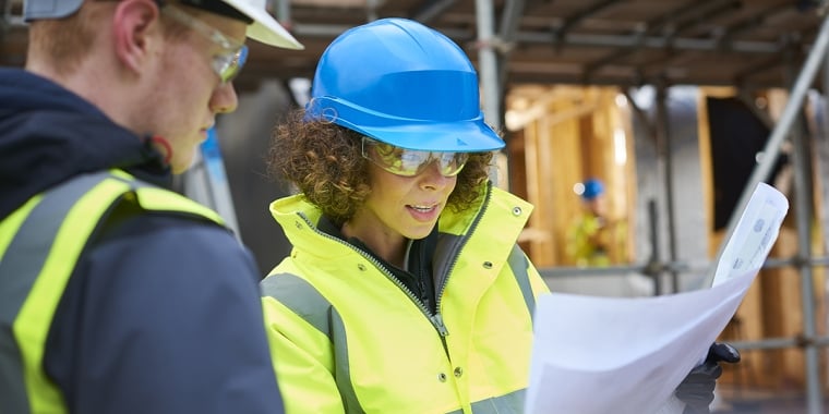 A female construction worker stands on a building site housing development and instructs a co-worker.