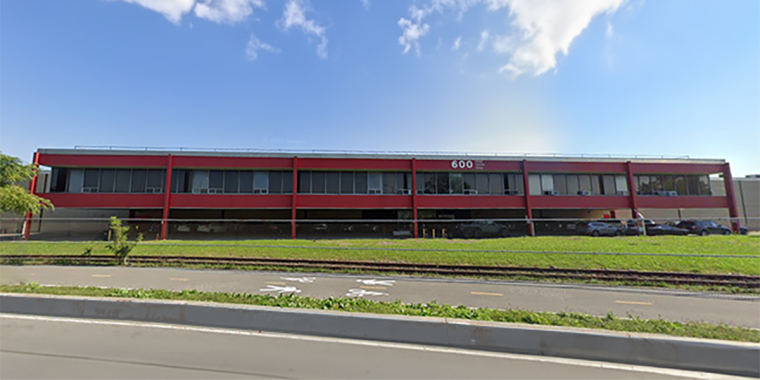 Exterior view of 600 Food Center Drive. A modern two-story red building with numerous windows, set against a clear blue sky with scattered clouds. A road and railroad tracks run parallel to it in the foreground. A few cars are parked beside the building.