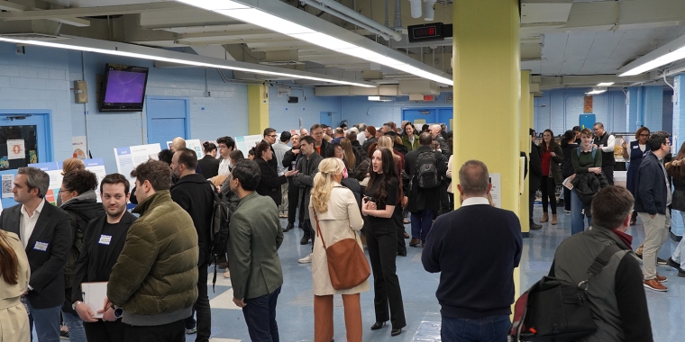 A large group of people gather in a room with blue walls and yellow pillars at a SPARC Community Engagement meeting. They appear to be engaged in conversations, some standing around tables with papers and displays. Overhead lights illuminate the space, creating a busy, social atmosphere.
