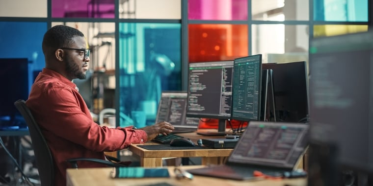 A Black man in a red shirt works at a desk with multiple computer monitors displaying code. The office has colorful glass panels in the background, adding a vibrant atmosphere to the modern workspace.
