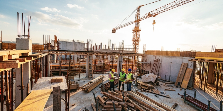 Three construction workers in safety gear discuss plans at a building site during sunset. Surrounding them are piles of wood, metal bars, and a crane, indicating ongoing construction. The skyline features a mix of clouds and blue sky.