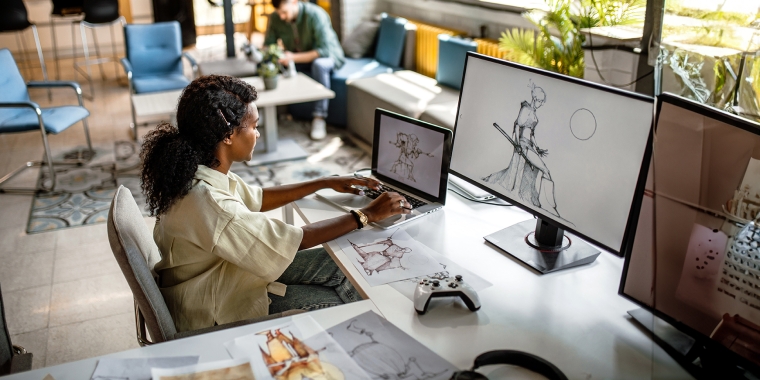 Young, black video game designer in company studio, working at her desk computer, drawing new sketches.