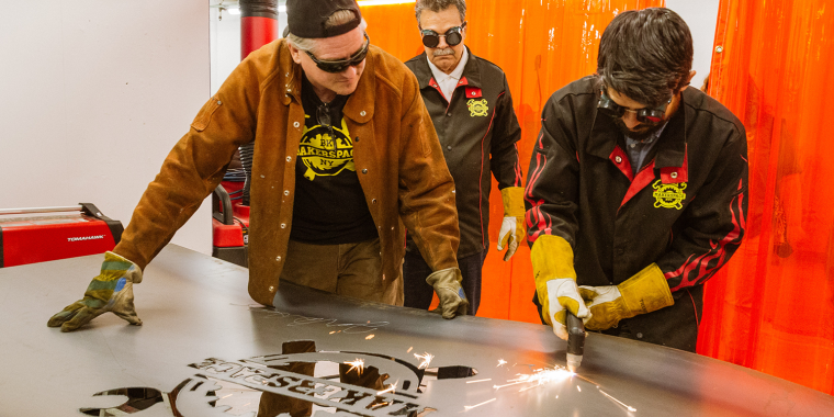 Three individuals in protective gear are working with metal, with one of them using a welding tool on a metallic surface.