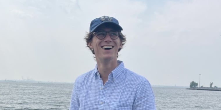 A man wearing a blue cap and glasses, Campbell Munn, smiling widely while standing near a body of water. He is dressed in a light blue, checkered long-sleeve shirt. The background shows a cloudy sky and distant shoreline.