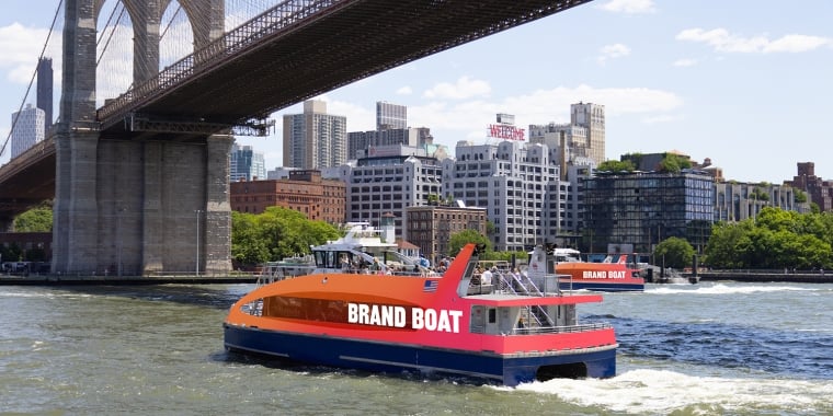 An orange and blue ferry labeled &quot;BRAND BOAT&quot; cruises under the Brooklyn Bridge with the New York City skyline in the background on a sunny day.