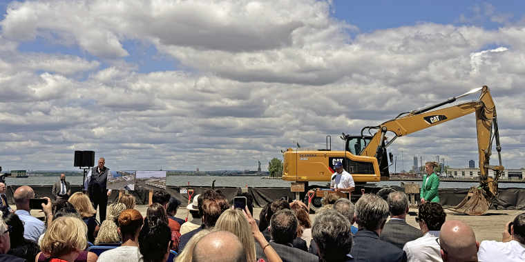 A group of people listening to NYC Mayor Eric Adams speaking at the South Brooklyn Marine Terminal groundbreaking ceremony with a large excavator in the background and a partly cloudy sky.