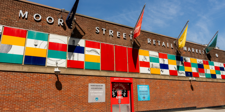 Exterior of Moore Street Retail Market, a brick building with colorful geometric panels and various flags. The entrance is marked by a red frame, flanked by informational signs. The sky is clear and blue.