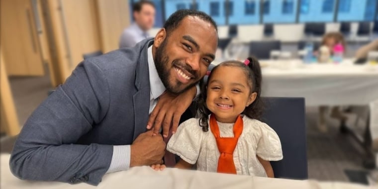 Justyn Turner in a suit, smiles while sitting next to a young girl wearing an oversized orange tie. They are seated at a table covered with white tablecloths in a brightly lit room with large windows.