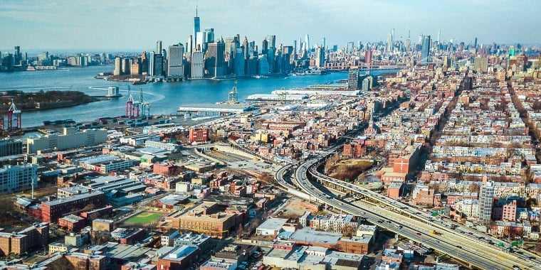 Aerial view of Red Hook Brooklyn featuring a mix of commercial, residential buildings, highways, and the New York Harbor with Manhattan skyscrapers in the background on a clear day.