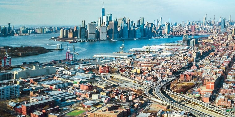 Aerial view of Red Hook Brooklyn featuring a mix of commercial, residential buildings, highways, and the New York Harbor with Manhattan skyscrapers in the background on a clear day.