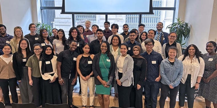 A group of summer interns pose for a photo in front of a screen displaying &quot;Blackstone LaunchPad.&quot; They are in an indoor setting with windows and plants in the background.