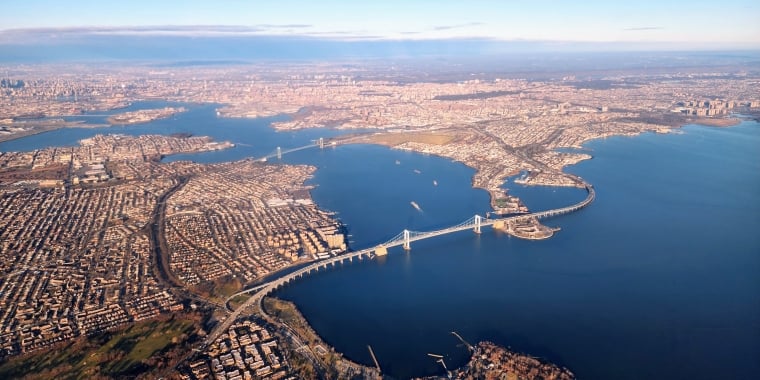 Aerial view of Flushing, Queens in NYC with a bridge spanning a large body of water, surrounded by densely packed buildings and green spaces under a clear blue sky.