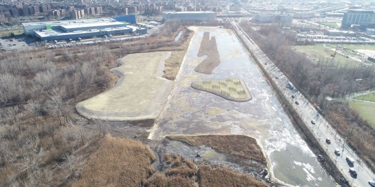 Aerial view of former Flushing Airport, a wetland area with surrounding urban landscape, showing water bodies, greenery, and a nearby road with moving cars.