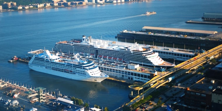 Aerial view of Manhattan cruise ships docked at Hudson River piers in Manhattan Cruise Terminal, with buildings and a waterfront in the background.