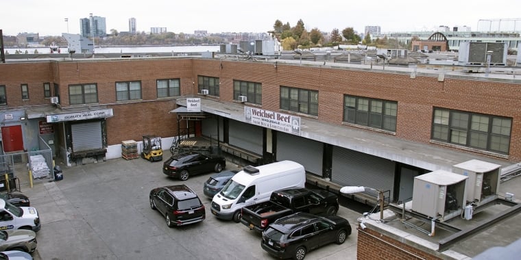 Aerial view of Gansevoort Square: A brick industrial building with loading docks, parked cars, and a sign reading &quot;Mitchell Beef.&quot; Urban landscape visible in the background.