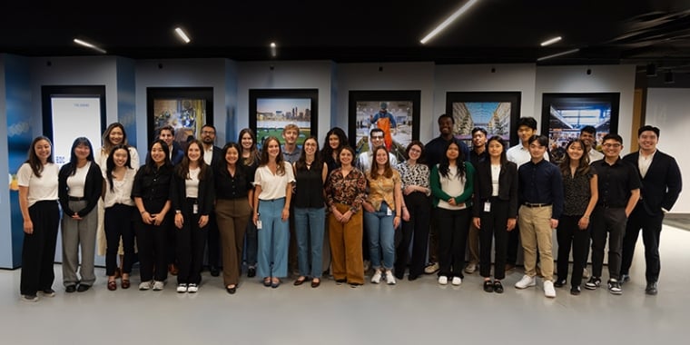 A group of people standing in a row indoors, posing for a photo. The background features framed artwork on the walls.