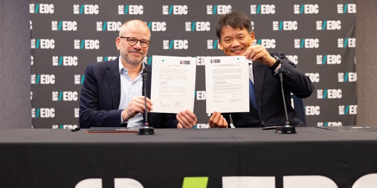 Two men seated at a table hold up signed MOU documents. A banner behind them displays the EDC logo repeatedly.