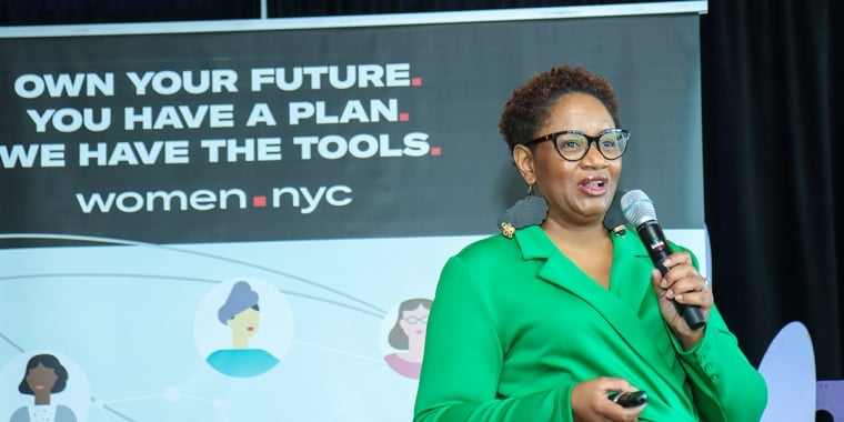 A person in a green blazer speaks into a microphone at an event with a sign reading "Own your future. You have a plan. We have the tools. women.nyc."