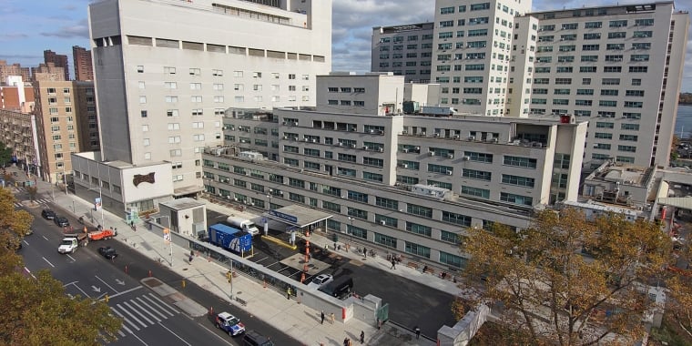 Aerial view of the large Metropolitan Hospital complex with multiple buildings and a busy street in the foreground. Trees with autumn foliage line the area.