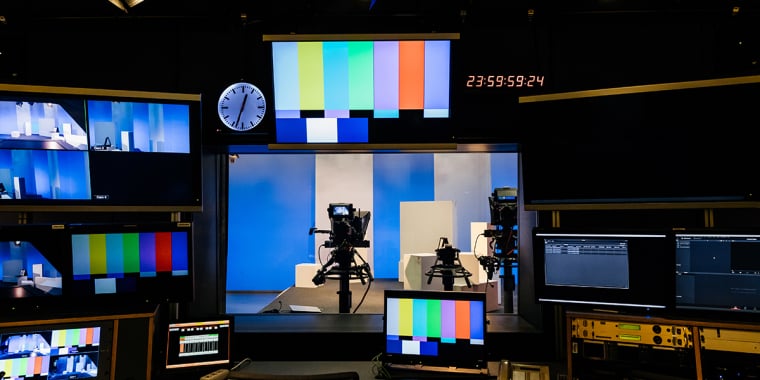 A television production control room with several monitors displaying test patterns and live feeds. A digital clock shows 23:59:58. Two cameras on tripods are in a studio setting visible through a window.