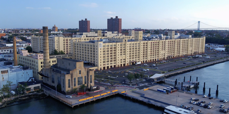 Aerial view of a waterfront industrial complex with a mix of modern and older buildings. A large factory-like structure is prominent, with residential high-rises in the background and a dock with vehicles in the foreground.