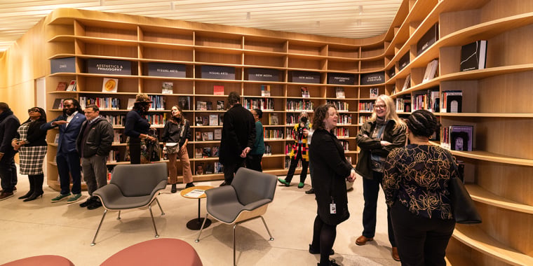 People are gathered in a modern library space with curved wooden shelves filled with books. Several small groups are conversing while others browse the selection. The area features comfortable seating and warm lighting.