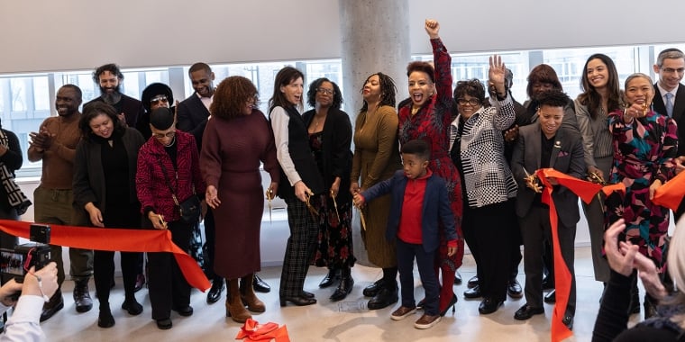 A diverse group of people celebrates at a ribbon-cutting ceremony inside a building. They hold a long red ribbon, and several individuals have their arms raised in excitement. Photographers are capturing the moment.