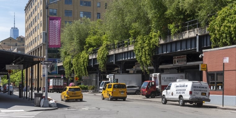 Street scene with a view of the High Line in New York City. The old railway track is lined with greenery and bordered by buildings. Yellow taxis, a van, and a café with a red awning are visible. The One World Trade Center is in the background.