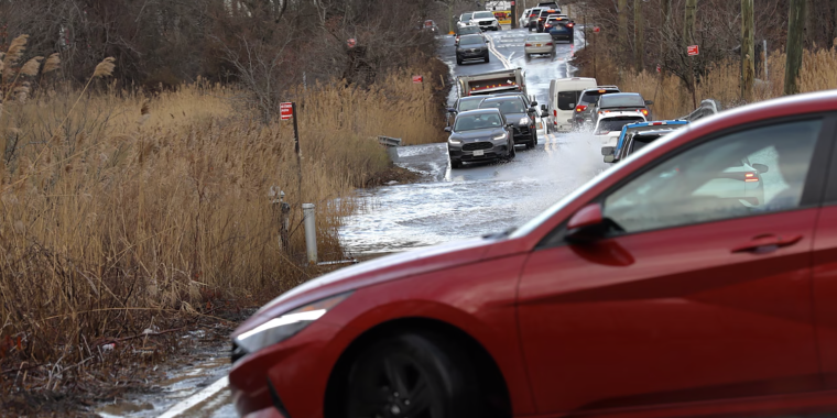 A red car crosses a flooded road, with other vehicles lined up behind it. The road is partially submerged, flanked by tall grass and trees. Overcast sky and power lines are visible in the background.