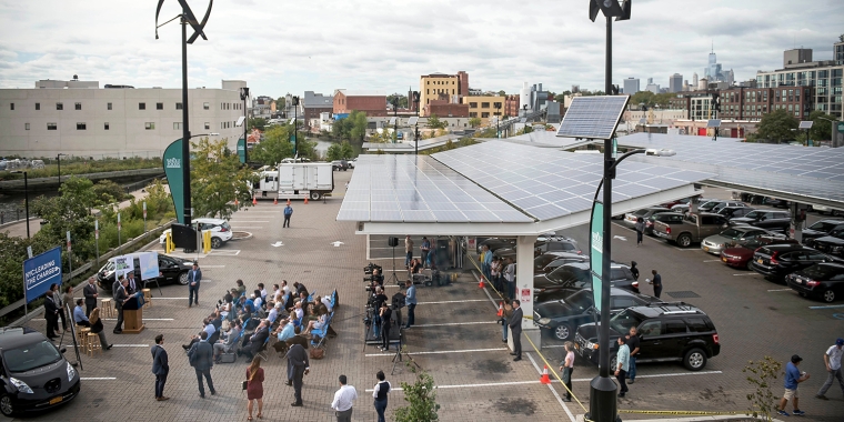 A parking lot features solar panels as a canopy. People are gathered for an event beneath the panels. Wind turbines are visible above, and there are banners and chairs set up. The background shows an urban landscape with buildings.