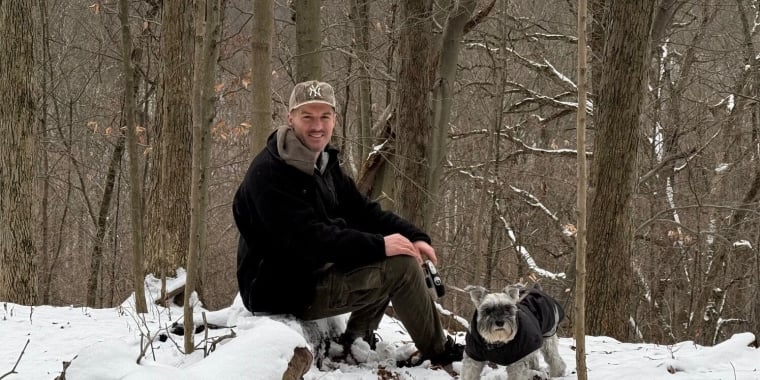 Matthew Furlong dressed in a black jacket and cap sits on a log in a snowy forest, holding a camera. Beside him is a small dog wearing a dark coat. Bare trees and a snowy landscape are in the background.