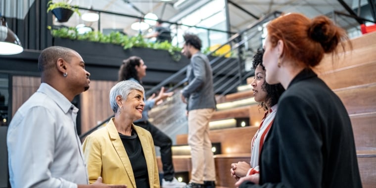A group of diverse people engage in conversation on wooden bleachers in a modern office space. Green plants adorn the upper levels. Some people are standing while others sit or ascend the staircase in the background.