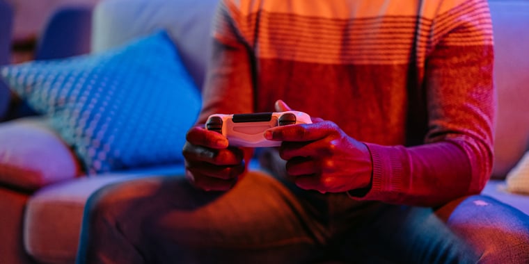 Person sitting on a couch, holding a video game controller. They are wearing a sweater and jeans. The room is dimly lit with colorful lights, and there are soft pillows and a small table with plants in the foreground.
