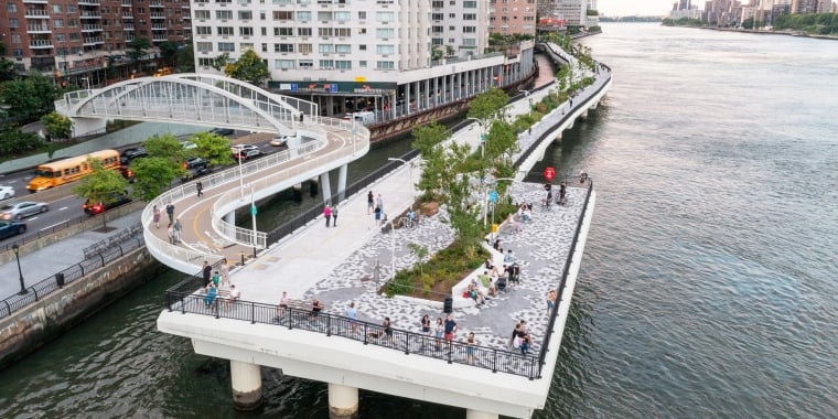 Aerial view of the East Midtown Greenway with a winding pedestrian bridge over the East River. People relax on benches surrounded by greenery. Adjacent is a city street with traffic, and tall buildings. A distant bridge is visible in the background.