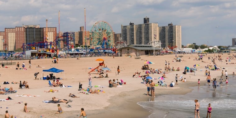 People relax on a sandy beach near the ocean, with colorful umbrellas, children playing in the water, and a cityscape with tall buildings, a Ferris wheel, and amusement park rides in the background.