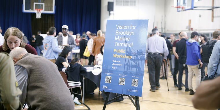 A group of people attend a public workshop in a gymnasium. A blue sign in the foreground reads Vision for Brooklyn Marine Terminal Public Workshop. Tables and informational materials are visible in the background.
