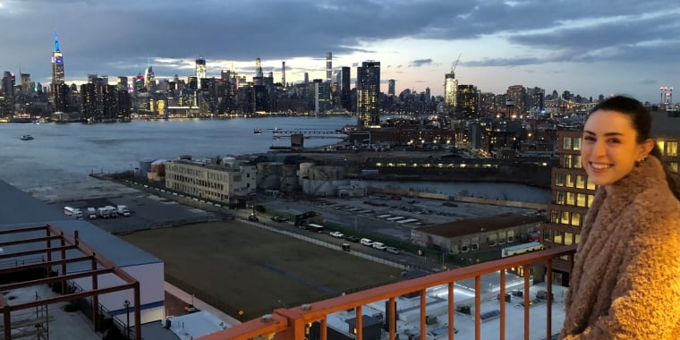 Mary Beth Williams in a brown coat stands on a balcony at sunset, smiling at the camera. Behind her is a New York City skyline with skyscrapers lit up and East River separating the city from the industrial buildings in the foreground.